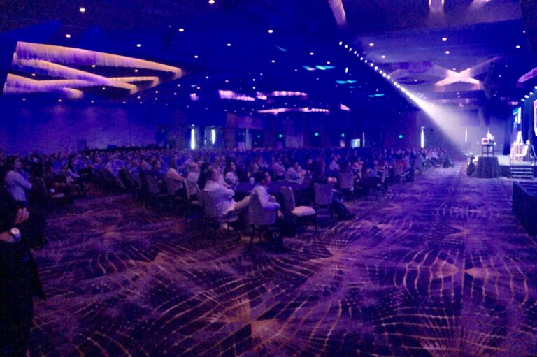 A conference hall audience under purple stage lights