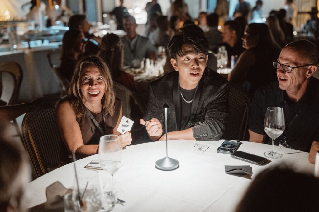 Johnny Wu showing a playing card to two smiling guests at a dinner table at a conference
